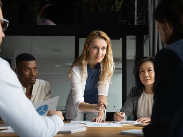 Confident skilled businesswoman explain marketing data paper report financial results analysis to multi-ethnic colleagues, company staff gathered at meeting brainstorming thinking strategizing concept